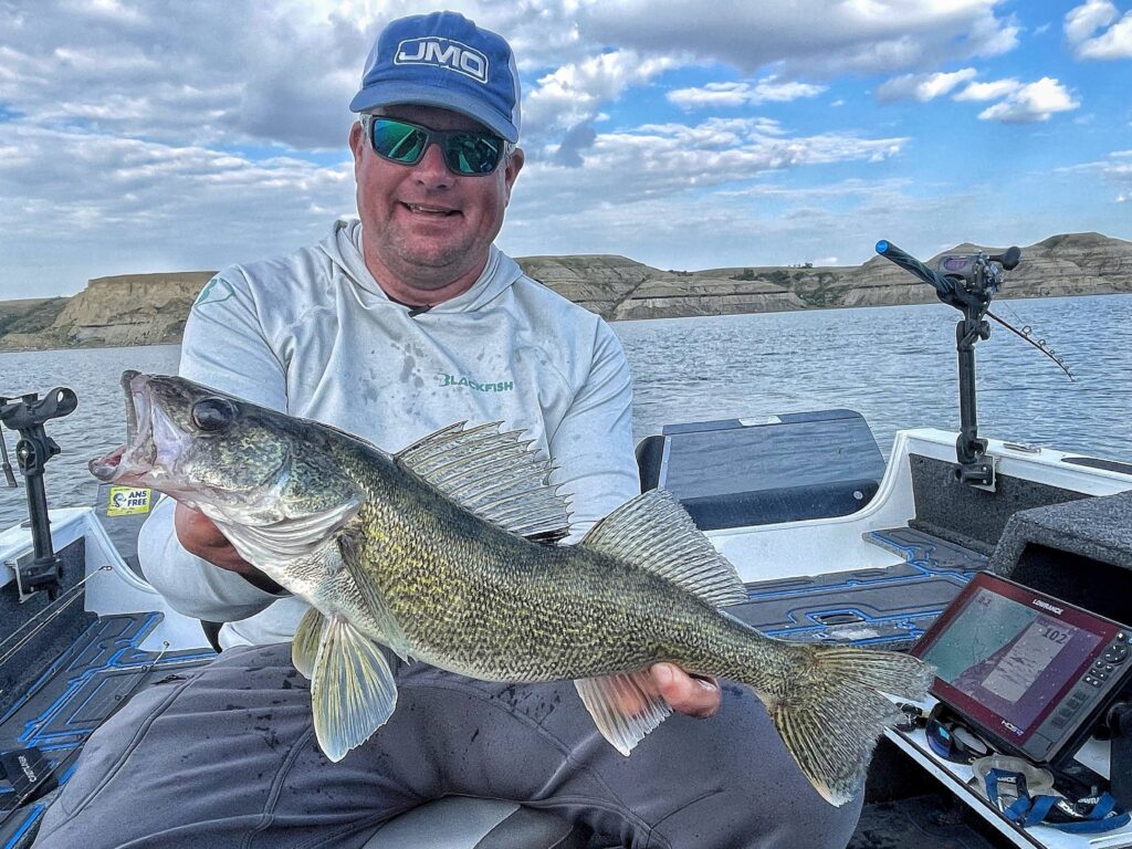 Lead Core and Small Cranks | Smiling angler wearing a blue JMO cap and sunglasses holds a large walleye fish aboard a boat on a scenic lake, with rolling hills and fishing gear in the background.