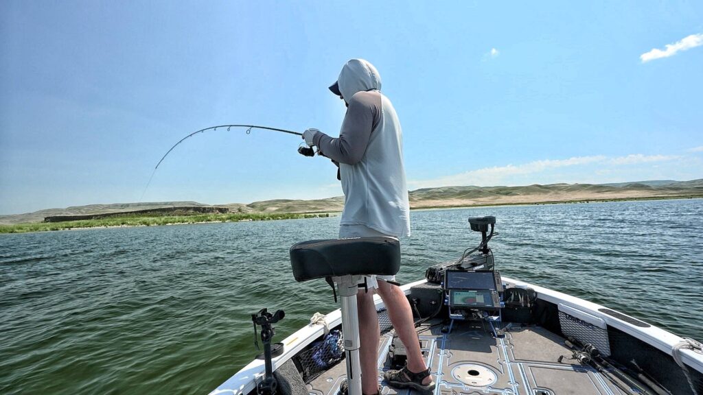 Pitchin’ Puppets | An angler reeling in a fish from a bass boat on a calm lake, with a scenic, hilly shoreline in the background.
